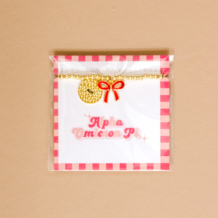 The Darling Effectโs Alpha Omicron Pi Bow Gold Charm Bracelet features gold beads, a red bow charm, and round pendant, displayed on an Alpha Omicron Pi card in pink gingham packagingโperfect for showing chapter pride.