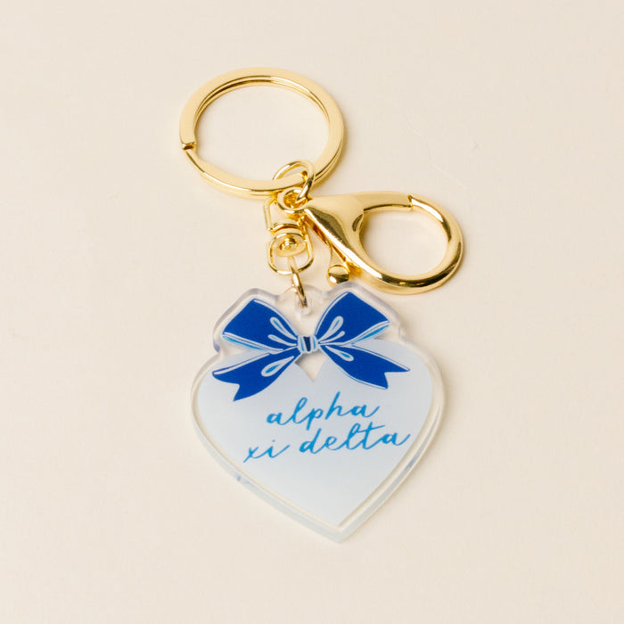 The Darling Effect Alpha Xi Delta Sweetheart Bow Acrylic Keychain features a heart shape, blue script, pastel bow, and gold keyring with claspโperfect as a sisterhood gift.