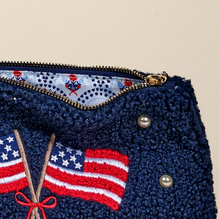 The Darling Effectโs American Flags Teddy Pouch is a navy textured sherpa pouch with embroidered flags, a red bow, pearl accents, and gold zipper, lined inside with white star and small red motif print fabric.