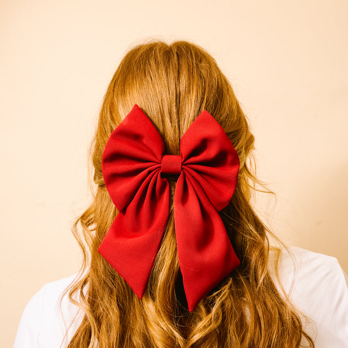 A person with long, wavy, reddish-brown hair is shown from behind, wearing The Darling Effectβs Burgundy Hair Bow as a bright accessory. The background is a solid light beige color.