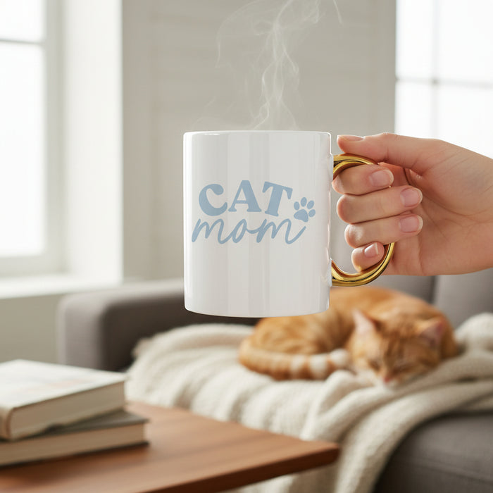 A hand holds The Darling Effectโs Cat Mom 11 oz Cozy Cute Ceramic Mug, featuring blue text and a paw print with a gold handle, as steam rises. In the background, an orange cat naps on a blanket-covered sofaโideal for coffee or tea moments.