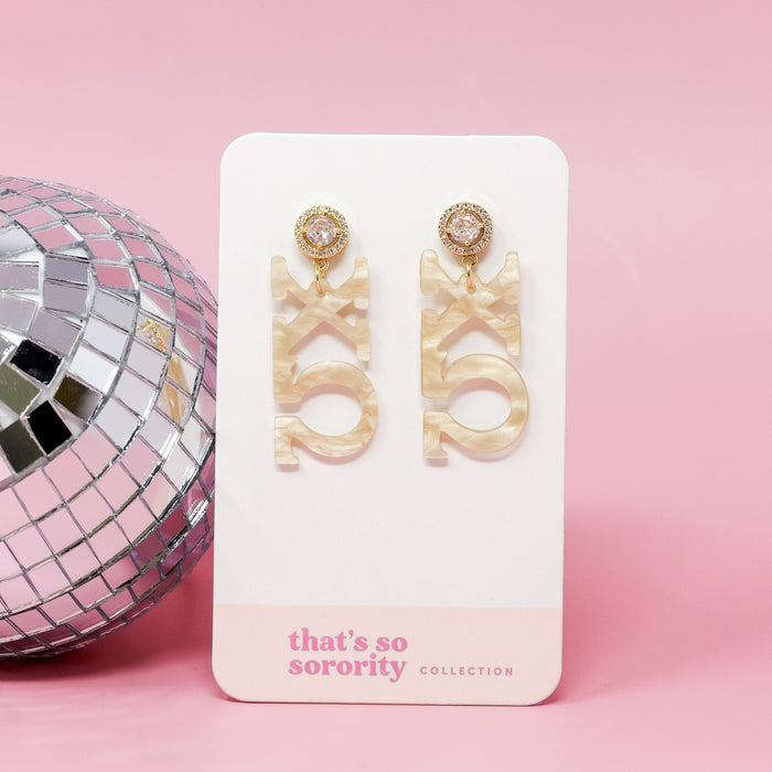 A pair of Chi Omega Letter Gold Acrylic Earrings by The Darling Effect hang on a white card labeled βthatβs so sorority collection,β set against a pink background with a silver disco ballβperfect for any bid-day basket.