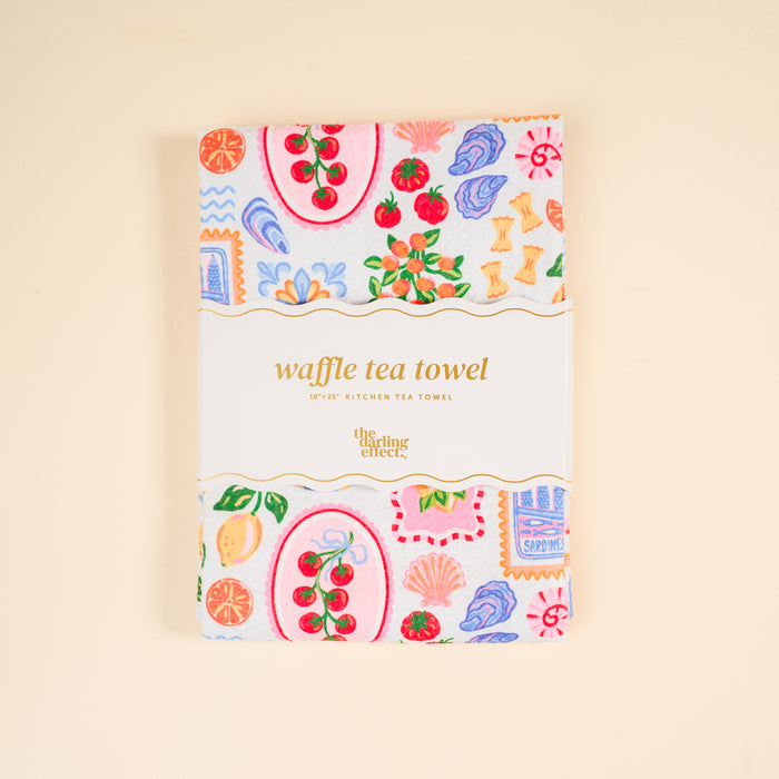A folded Ciao Bella Waffle Tea Towel by The Darling Effect, featuring colorful fruit and floral patterns and a label, sits on a beige background. This absorbent 18x28" kitchen towel adds charm to any kitchen.