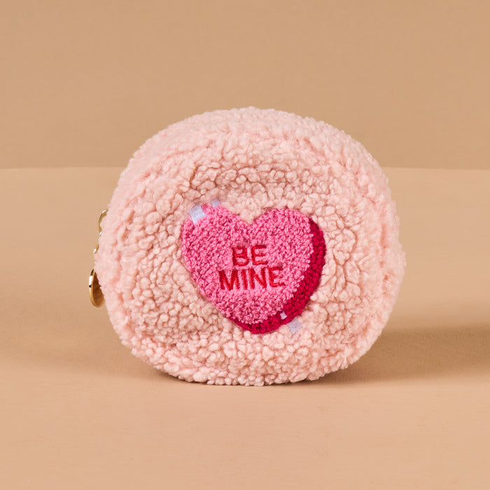 The Darling Effect Circle Be Mine Teddy Pouch is a small, round pink sherpa pouch with a pink heart and “BE MINE” in red embroidery—perfect for cosmetics or gifting. Shown against a plain beige background.