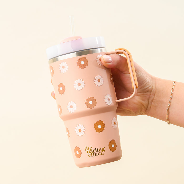 A hand holds The Darling Effect's Darling Daisy Tan 20 oz Mini On-The-Go tumbler, decorated with white and brown daisies, a straw, and "the darling effect" in gold near the bottom.