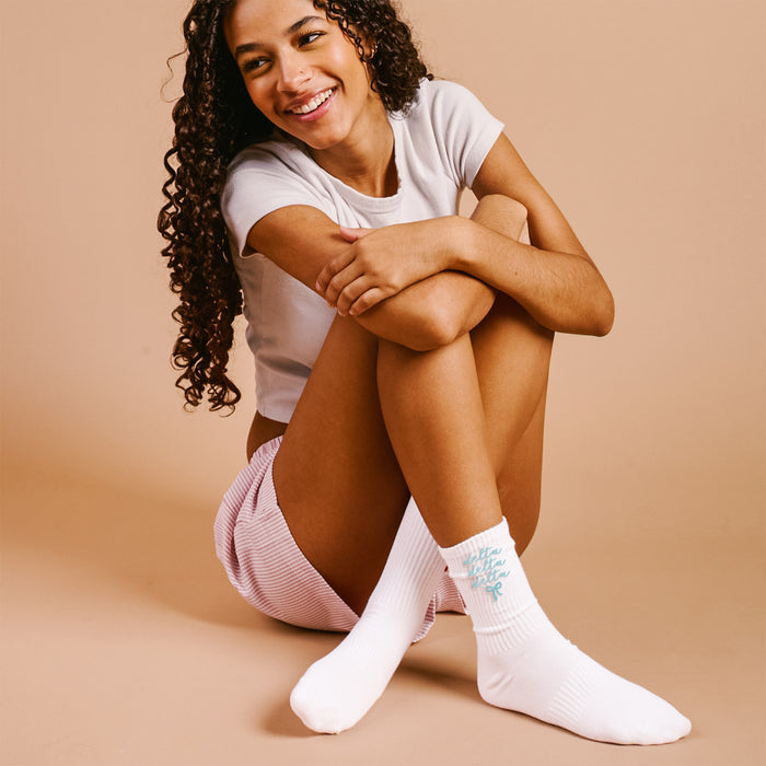 A young woman with curly hair sits on the floor, smiling, in a white t-shirt, pink striped shorts, and The Darling Effect’s Delta Delta Delta Embroidered Crew Socks—perfect sorority gifts—against a beige background.