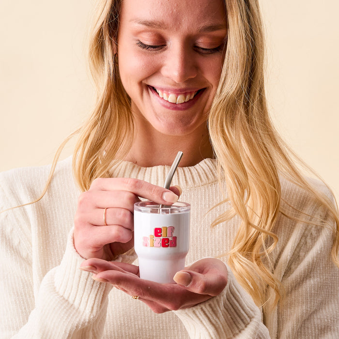 A woman with long blonde hair stirs the-darling-effect's Elf Sized 2 oz Tiny Tumbler with a metal straw, wearing a cream sweater against a neutral background.