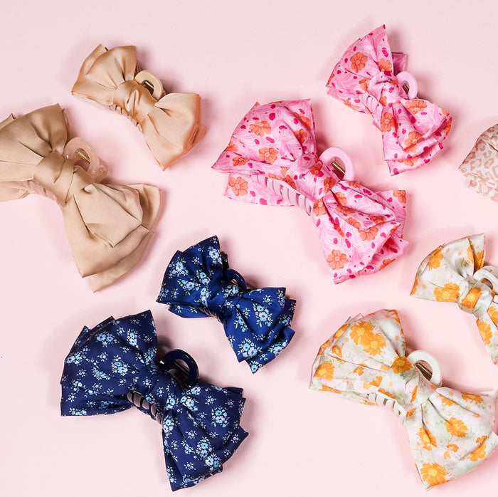 The Endless Daydream Cream Satin Bow Claw Clip by the-darling-effect is displayed alongside bows in beige, pink floral, navy floral, and white-orange bloomsβcreating a charming accessory set on a pale pink background.
