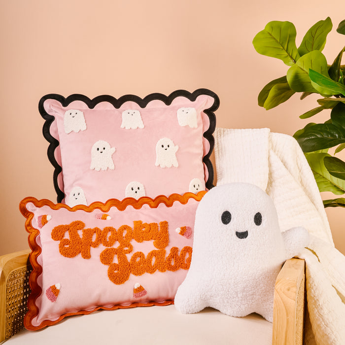 A cozy chair with a white blanket holds The Darling Effect Boo Crew Scalloped Square Throw Pillowβpink with small ghostsβplus an orange βSpooky Sznβ pillow and plush ghost, next to a leafy plant against a beige background.
