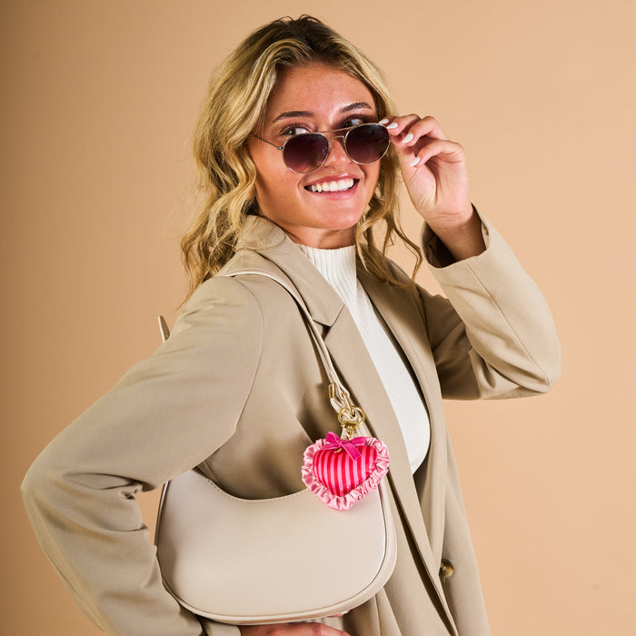 A woman in a beige blazer, white top, and sunglasses smiles while holding her sunglasses. She carries a cream handbag adorned with The Darling Effectβs Heartthrob Puffy Heart Bag Charm, standing against a beige background.