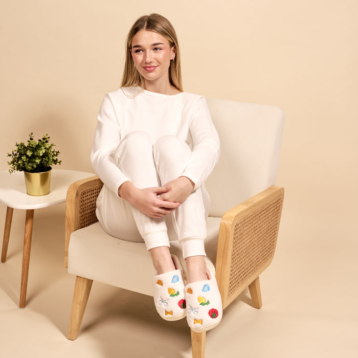A young woman in white loungewear sits cross-legged on a beige armchair, smiling softly. She wears The Darling Effectβs Italian Summer Fuzzy Slippers with colorful fruit designs. A small potted plant rests on the side table beside her.