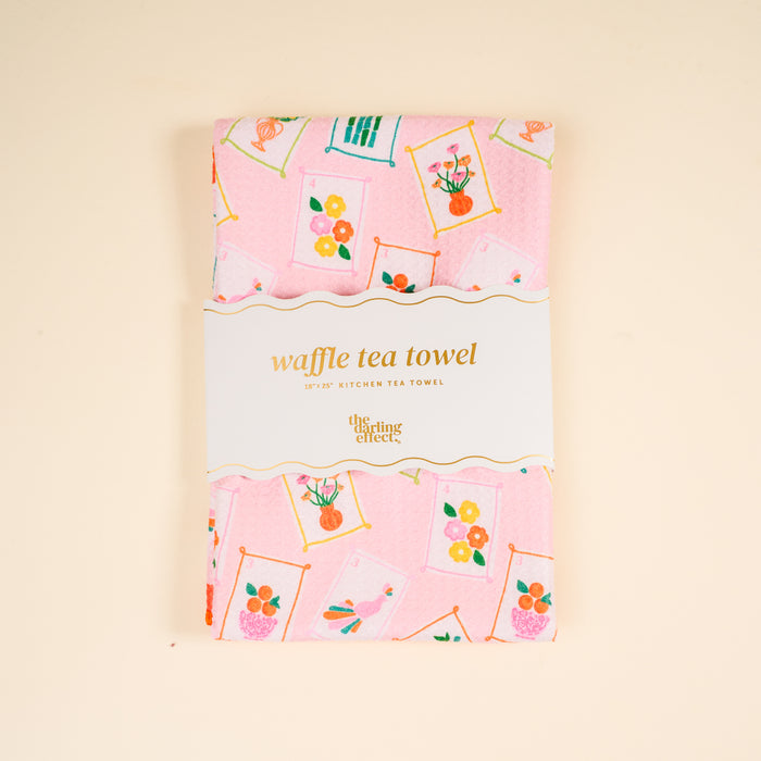 A pink decorative tea towel with framed flower and bird prints, folded and wrapped with a label that reads "Itβs Mahj Time Waffle Tea Towel" from The Darling Effect. Absorbent and perfect for everyday kitchen use.