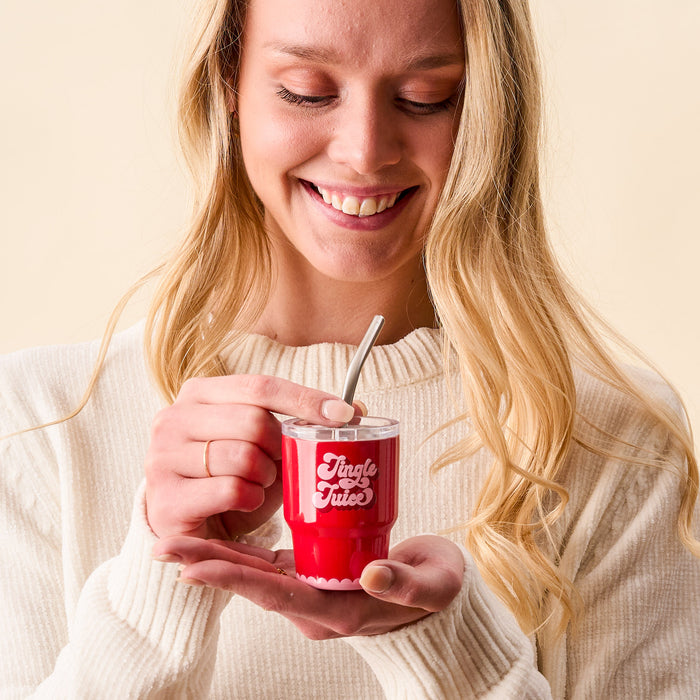 A smiling woman in a cream sweater holds the-darling-effectβs Jingle Juice 2 oz Tiny Tumbler with a metal straw, looking down at her drink against a neutral background.