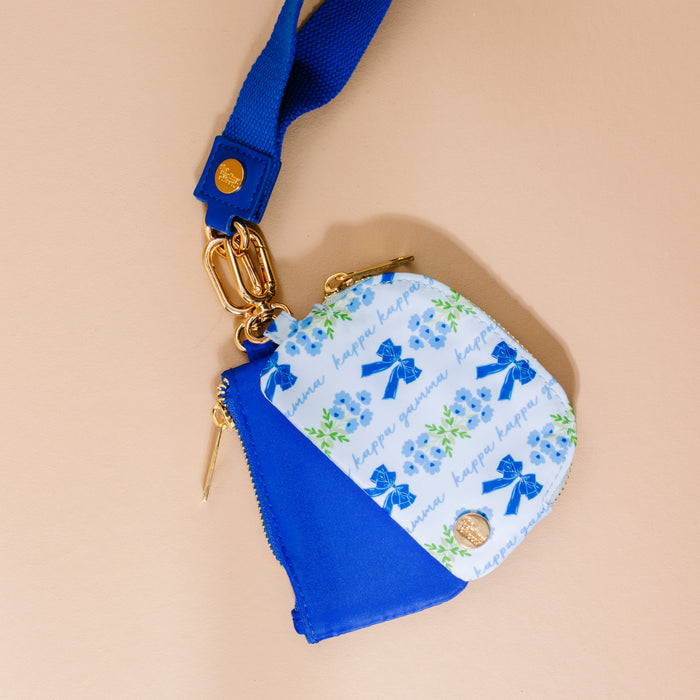 The Darling Effect Kappa Kappa Gamma Dynamic Duo Pouch Wristlet features a blue strap, gold hardware, and a blue-and-white floral pattern with "happy guava" and blue bows, shown on a beige surface.