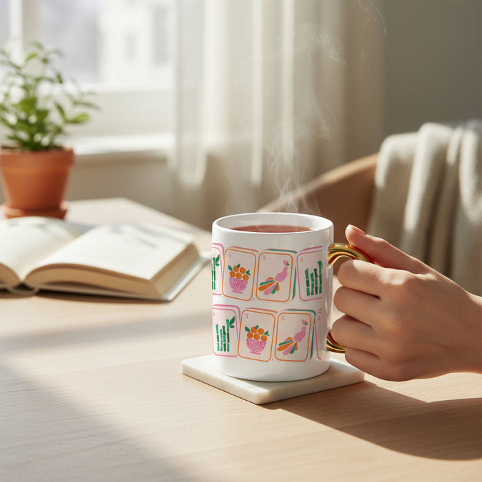 A hand holds The Darling Effectโs Mahjong Tiles 11 oz Cozy Cute Ceramic Mug, featuring colorful tile designs and a gold handle, resting on a coaster beside an open book, potted plant, and soft fabric on a sunlit wooden table.
