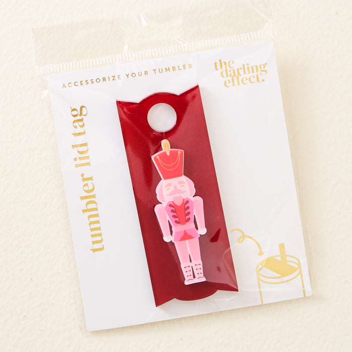 The Nutcracker Tumbler Lid Tag by The Darling Effect features a pink nutcracker on a red background with gold "tumbler lid tags" and "the darling effect" lettering. Fits Stanley Quencher 2.0βperfect for holiday tumbler accessories.