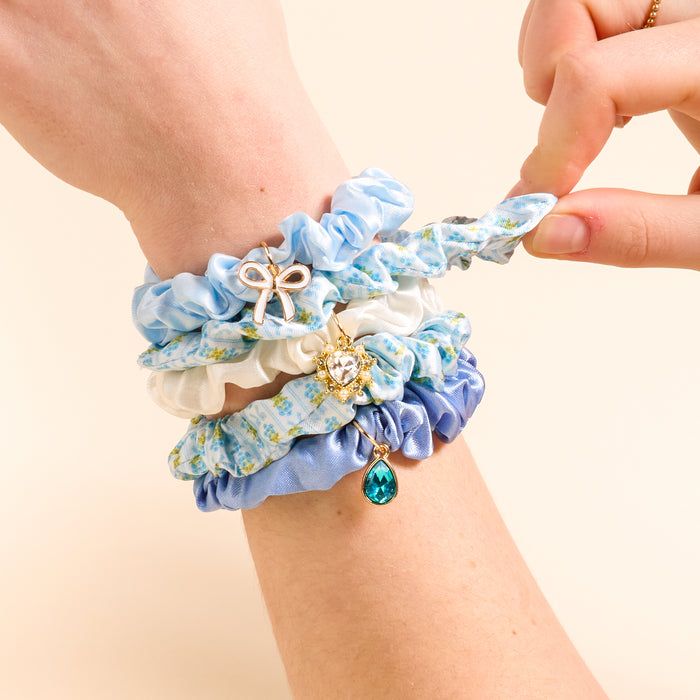 A hand displays the Oh So Fancy Blue Scrunchie Charm Set by The Darling Effectβblue and white floral scrunchies adorned with gold charms, including a bow, crystal heart, and teal teardropβagainst a beige background.