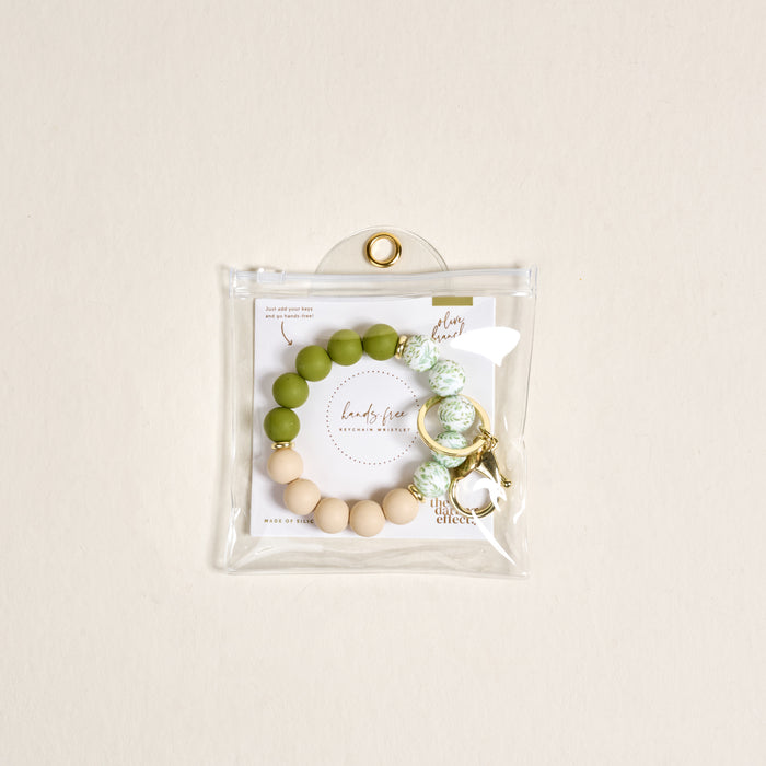 The Darling Effectβs Olive Branch Hands-Free Keychain Wristlet features green, white, and beige silicone beads with a gold keyring, packaged in a clear snap pouch and includes a branded card for extra style.