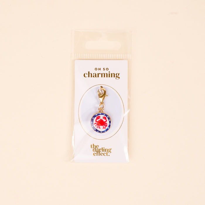 The Darling Effect Painted Crab Jewelry Charm Gold features a red crab on a blue and white background with a gold clasp, perfect for your charm bar. Packaged on a white Oh So Charming card, itโs a delightful jewelry addition.