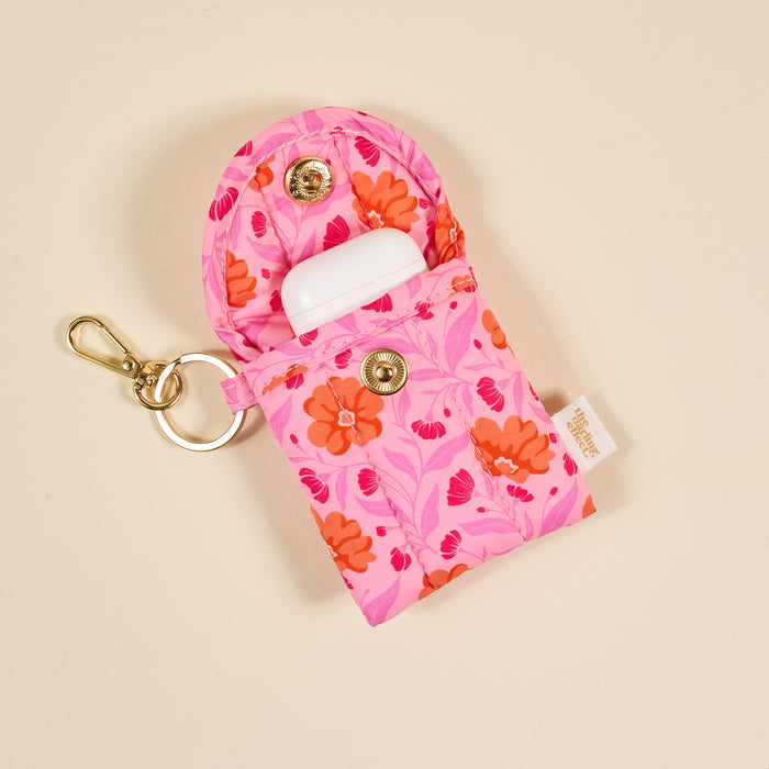 The Darling Effectโs Poppin' Petals Hot Pink Puffy Earbud Case features a pink floral design, snap button closure, gold keyring and clasp, white wireless earbuds inside, and a side fabric tag for a stylish keychain accessory.