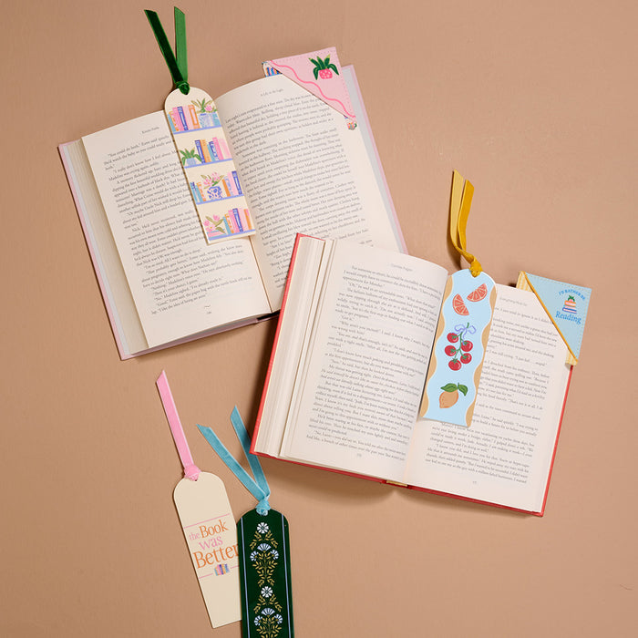 Flat lay of three open books on a tan surface, each with a Book Shelfie Acrylic Bookmark. Three additional bookmarksβwith designs featuring books, plants, text, fruit, and velvet ribbon tasselsβare arranged around them. Perfect for book lovers!.