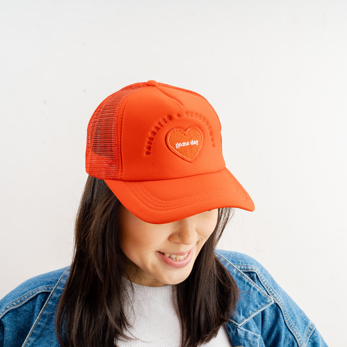 A woman with long dark hair smiles as she looks down, wearing The Darling Effectβs Red Game Day Trucker Hat with an embroidered heart and mesh adjustable back. She pairs it with a white shirt and blue denim jacket against a plain background.