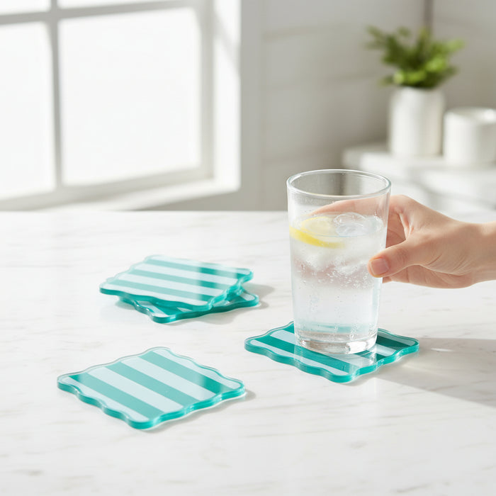 A hand holds a glass of water with lemon on a white marble table, next to The Darling Effect’s Sicily Stripes Teal Table Talk Acrylic Coaster Set. Sunlight streams in, and a potted plant adds fresh décor in the background.