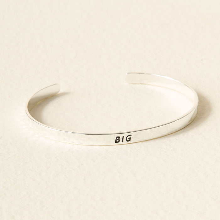 The Darling Effectβs Silver Big Sorority Cuff features βBIGβ engraved in black on a simple silver band, shown on a light, textured surface.