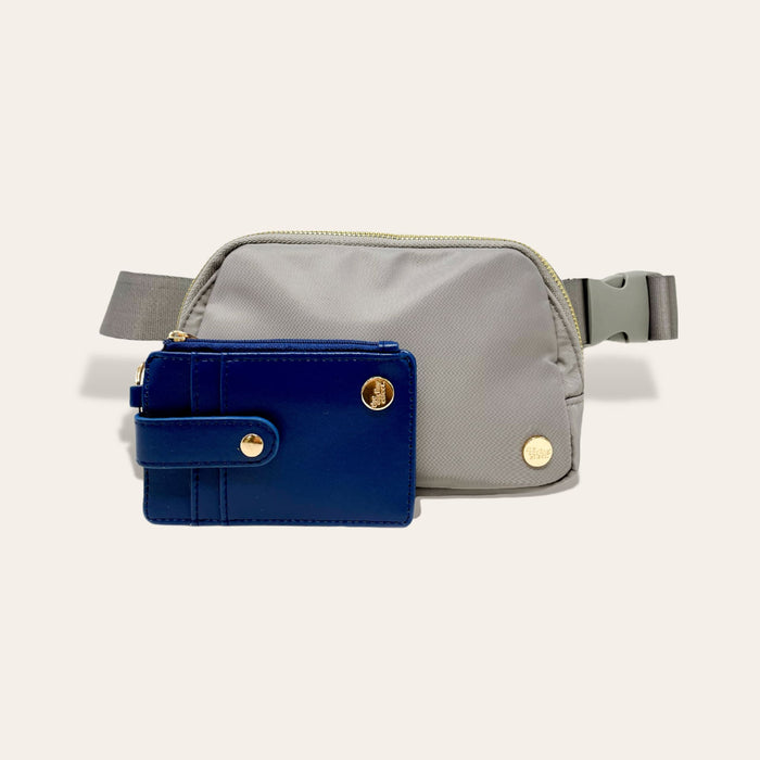 The Darling Effect's Slate Gray Belt Bag with zipper and adjustable strap is placed behind a matching wallet with gold button accents, both displayed on a plain off-white background.