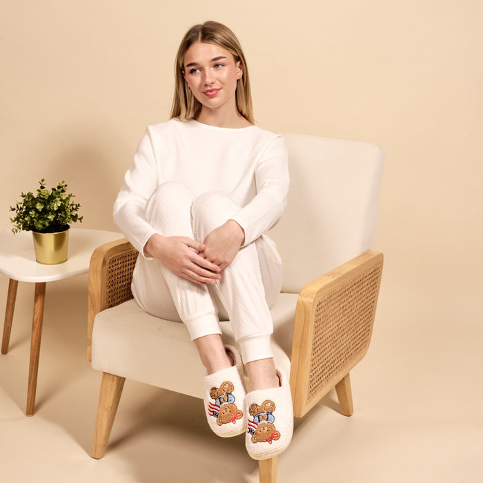 A young woman in white loungewear sits on a beige armchair, legs crossed, wearing The Darling Effectβs Star Spangled Bear Fuzzy Slippers. A potted plant rests on a nearby small table against a soothing light beige background.