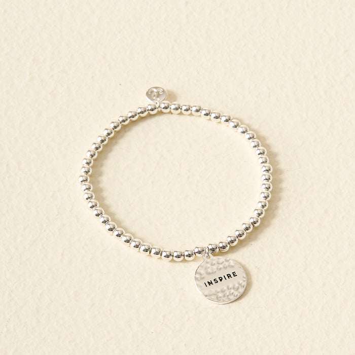 The Darling Effect's Stay Inspired Beaded Charm Bracelet features silver beads and a round charm engraved with "INSPIRE," making it a thoughtful gift. Shown on a cream-colored textured background.