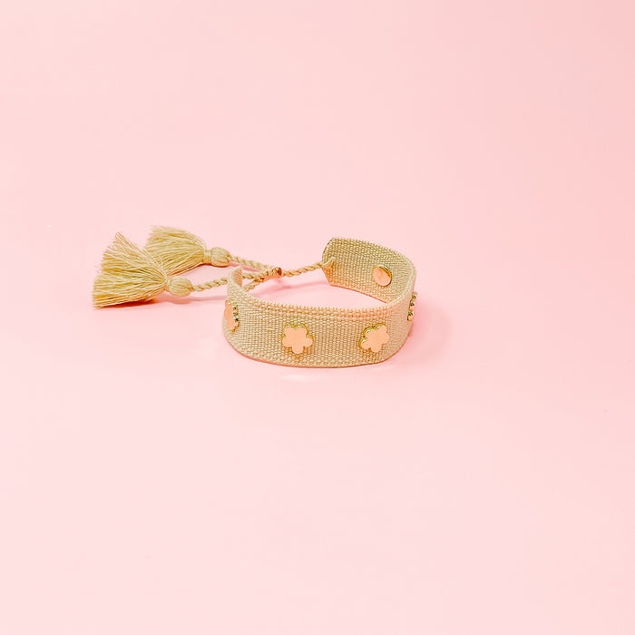 The Darling Effect Tan Woven Bracelet features gold clover-shaped accents and two tassels, shown on a light pink background.