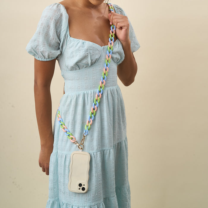 A woman in a light blue dress models The Darling Effectβs Tri Lively Hold the Phone Crossbody Chain, featuring a pastel multicolored strap and wavy-edged white holder, adding style against a beige backdrop.