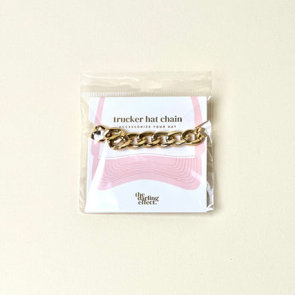 The Darling Effect Acrylic Link Gold Trucker Hat Chain shines on a light background and is packaged on a card with pink hat graphics and "the darling effect" at the bottom.