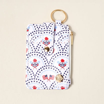 The Darling Effect’s All-American Darling Keychain Wallet features a keyring, card holder, snap closure, and zip pocket with gold hardware. Decorated with blue stars, red flowers, and American flag prints on a white background.
