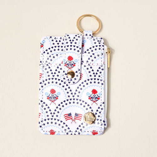 The Darling Effect’s All-American Darling Keychain Wallet features a keyring, card holder, snap closure, and zip pocket with gold hardware. Decorated with blue stars, red flowers, and American flag prints on a white background.