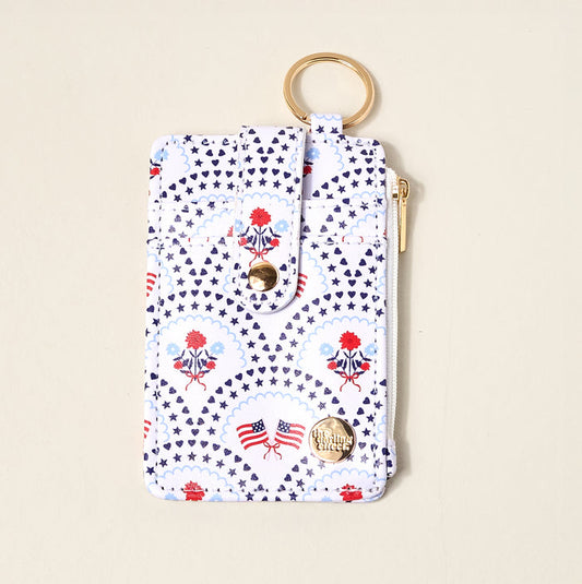 The Darling Effect’s All-American Darling Keychain Wallet features a keyring, card holder, snap closure, and zip pocket with gold hardware. Decorated with blue stars, red flowers, and American flag prints on a white background.