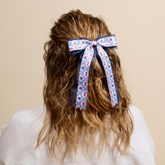 A person with wavy, light brown hair styled half-up and tied with The Darling Effect’s All-American Darling Lovely Layered Hair Bow is shown from behind in a white textured top against a beige background, highlighting their chic taste in accessories.