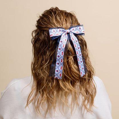 A person with wavy, light brown hair styled half-up and tied with The Darling Effect’s All-American Darling Lovely Layered Hair Bow is shown from behind in a white textured top against a beige background, highlighting their chic taste in accessories.