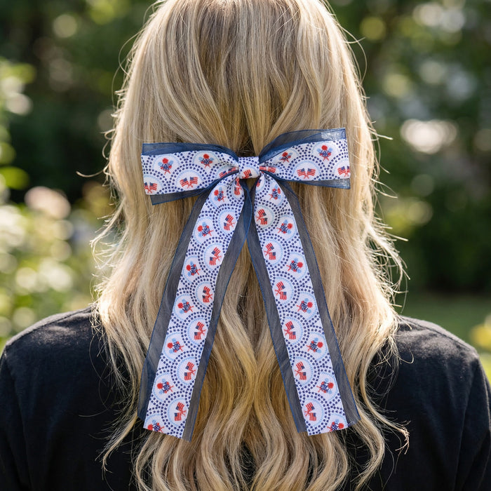 Wearing The Darling Effect's All-American Darling Lovely Layered Hair Bow, a person with long, wavy blonde hair stands outdoors with their back to the camera. The bold blue, white, and red accessory makes an eye-catching statement.
