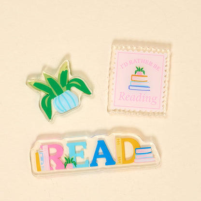 The Darling Effect's All Booked Up Little Magnet Set features three charming acrylic magnets—a plant, a "I'd rather be reading" stamp, and colorful READ letters—perfect fridge decor for any book lover.