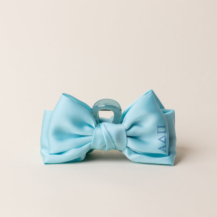 The Darling Effect’s Alpha Delta Pi Satin Bow Claw Clip features a light blue satin bow with ΑΔΠ Greek letters on the right, attached to a translucent light blue claw clip—perfect for updos and shown on an off-white background.