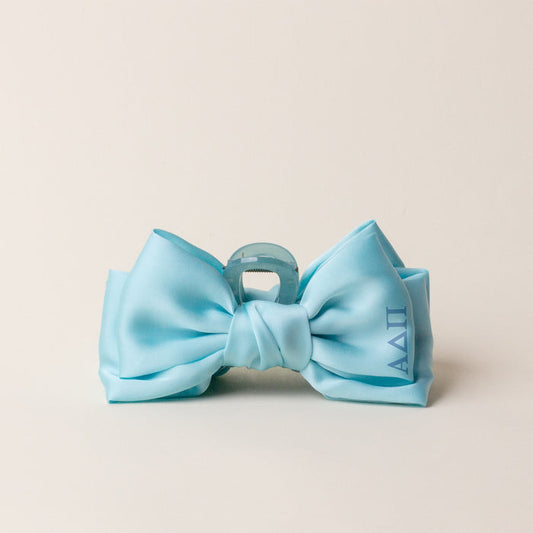 The Darling Effect’s Alpha Delta Pi Satin Bow Claw Clip features a light blue satin bow with ΑΔΠ Greek letters on the right, attached to a translucent light blue claw clip—perfect for updos and shown on an off-white background.