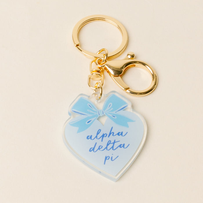 The Darling Effect's Alpha Delta Pi Sweetheart Bow Acrylic Keychain features a gold lobster clasp, a heart-shaped charm with a pastel blue bow, and “alpha delta pi” in blue script—an adorable sisterhood gift.