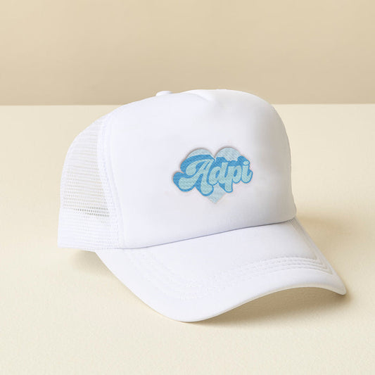 The Darling Effect Alpha Delta Pi White Trucker Hat features white mesh and a heart-shaped patch with "Adpi" in light blue retro-style letters, embroidered on the front. Displayed on a light surface with a beige background.