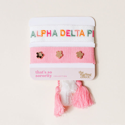 The Darling Effect's Alpha Delta Pi Woven Bracelet Set includes two pink and white bracelets on a card—one with ALPHA DELTA PI in colorful letters, the other with gold flower studs—plus matching tassels, perfect for sorority gifting.