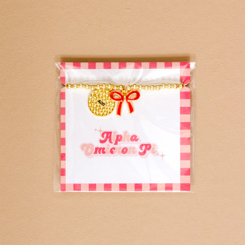 The Darling Effect’s Alpha Omicron Pi Bow Gold Charm Bracelet features gold beads, a red bow charm, and round pendant, displayed on an Alpha Omicron Pi card in pink gingham packaging—perfect for showing chapter pride.
