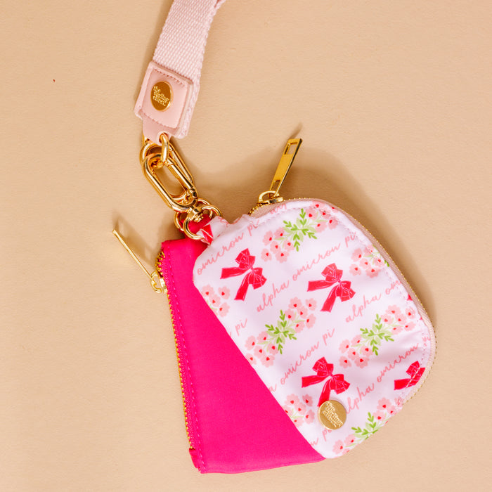 The Darling Effect Alpha Omicron Pi Dynamic Duo Pouch Wristlet features a pink and white floral design with red bows, sorority text, a light pink strap, and gold hardware—combining style and practicality.