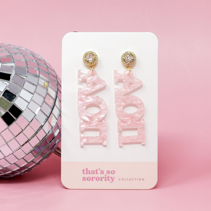 A pair of Alpha Omicron Pi Letter Pink Acrylic Earrings by The Darling Effect, displayed on a “that’s so sorority” card beside a silver disco ball, make the perfect bid-day basket gift, all set on a light pink background.