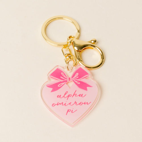The Darling Effect’s Alpha Omicron Pi Sweetheart Bow Acrylic Keychain features a gold keyring with a pink heart charm, pastel bow, and “Alpha Omicron Pi” in cursive—perfect as a sisterhood gift.